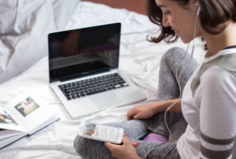 A woman sitting on her bed with an open laptop and reading an article on her phone.