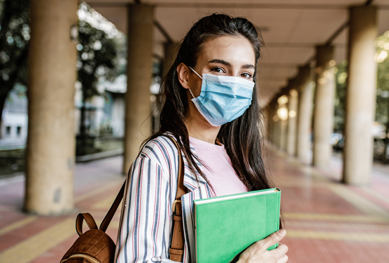 A college student standing outside a college campus wearing a mask and holding a book.