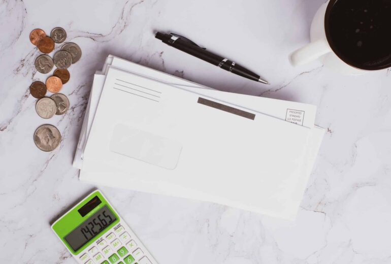 Desk scene with coins scattered beside envelopes, a black pen, a coffee cup, and a green calculator on a white marble surface.